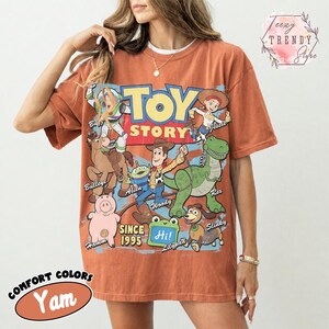 May include: A rust-colored short-sleeve t-shirt featuring a large graphic print of Toy Story characters including Woody, Buzz Lightyear, Jessie, Rex, Hamm, Bullseye, Slinky Dog, and an Alien. The text reads Toy Story Since 1995 and Hi.
