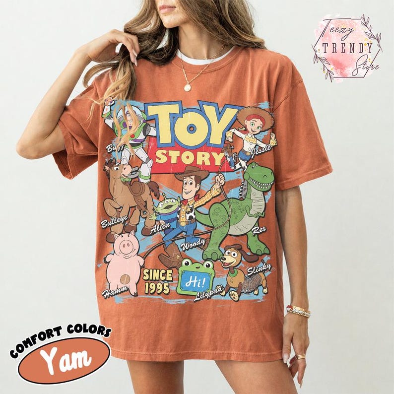 May include: A rust-colored short-sleeve t-shirt featuring a large graphic print of Toy Story characters including Woody, Buzz Lightyear, Jessie, Rex, Hamm, Bullseye, Slinky Dog, and an Alien. The text reads Toy Story Since 1995 and Hi.