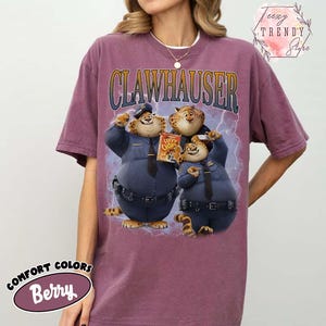 Puede incluir: Camiseta de color bayas con la palabra "CLAWHAUSER" sobre un gr&aacute;fico de tres personajes de dibujos animados con uniformes de polic&iacute;a. Los personajes sostienen una caja de cereales Lucky Chunks. La camiseta tiene una etiqueta Comfort Colors.