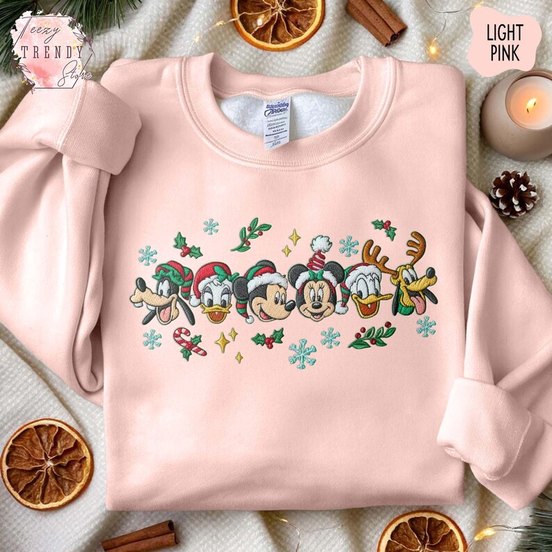 May include: Light pink sweatshirt featuring embroidered Disney characters in Christmas hats and decorations. The design includes Mickey Mouse, Minnie Mouse, Donald Duck, Goofy, and Pluto. The sweatshirt has a crew neck and long sleeves.