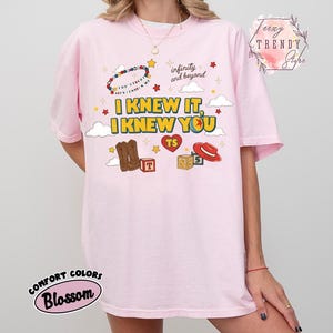 Puede incluir: Una camiseta rosa claro con un estampado gr&aacute;fico que incluye el texto I KNEW IT, I KNEW YOU, infinity and beyond, y YOU'VE GOT A FRIEND IN ME. El dise&ntilde;o presenta ilustraciones de botas vaqueras, un sombrero rojo, bloques y un coraz&oacute;n.