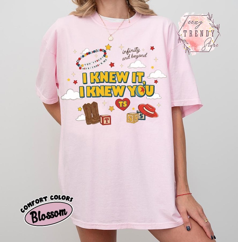 Puede incluir: Una camiseta rosa claro con un estampado gr&aacute;fico que incluye el texto I KNEW IT, I KNEW YOU, infinity and beyond, y YOU'VE GOT A FRIEND IN ME. El dise&ntilde;o presenta ilustraciones de botas vaqueras, un sombrero rojo, bloques y un coraz&oacute;n.