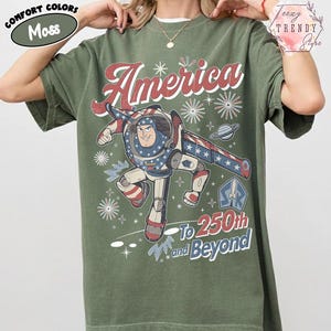 Puede incluir: Una camiseta de manga corta color verde musgo con un gr&aacute;fico de un personaje de ranger espacial con traje rojo, blanco y azul. El texto dice America, To 250th and Beyond, acompa&ntilde;ado de ilustraciones de fuegos artificiales, estrellas y un planeta.