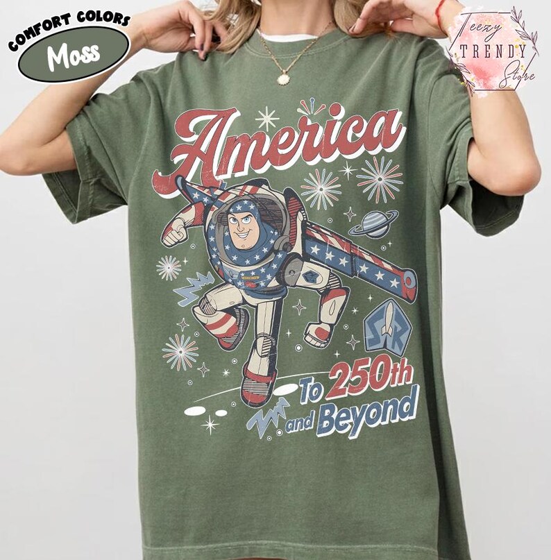 Puede incluir: Una camiseta de manga corta color verde musgo con un gr&aacute;fico de un personaje de ranger espacial con traje rojo, blanco y azul. El texto dice America, To 250th and Beyond, acompa&ntilde;ado de ilustraciones de fuegos artificiales, estrellas y un planeta.