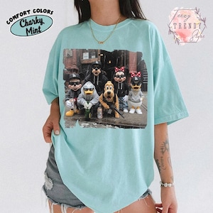 May include: A light mint green t-shirt featuring a graphic print of six cartoon characters wearing streetwear, including hoodies, baseball caps, and sunglasses. The characters are posed together in an urban setting with various accessories.