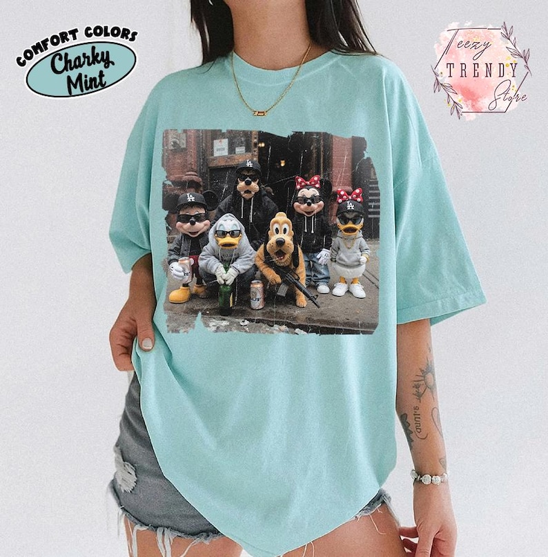 May include: A light mint green t-shirt featuring a graphic print of six cartoon characters wearing streetwear, including hoodies, baseball caps, and sunglasses. The characters are posed together in an urban setting with various accessories.