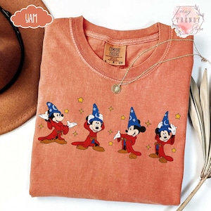 May include: Coral-colored t-shirt featuring a graphic of Mickey Mouse in a sorcerer's outfit with a blue hat and red robe. The design includes yellow stars. The shirt has a crew neck and a Comfort Colors tag.