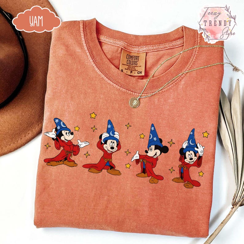 May include: Coral-colored t-shirt featuring a graphic of Mickey Mouse in a sorcerer's outfit with a blue hat and red robe. The design includes yellow stars. The shirt has a crew neck and a Comfort Colors tag.