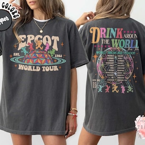 May include: Dark gray t-shirt featuring a colorful graphic of EPCOT World Tour with the text "EPCOT WORLD TOUR" and "EST. 1982". The back of the shirt has the text "DRINK AROUND THE WORLD" and a list of countries.