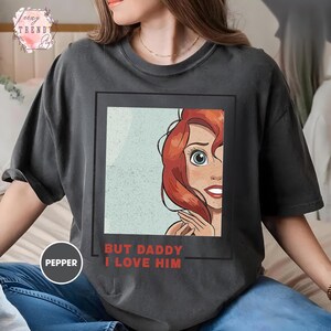 Puede incluir: Camiseta gris oscuro con una ilustraci&oacute;n enmarcada de un personaje de dibujos animados con pelo rojo y ojos azules. El texto rojo "BUT DADDY I LOVE HIM" est&aacute; impreso debajo. La camiseta tiene un corte holgado.