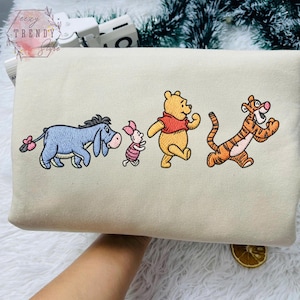 May include: Cream-coloured sweatshirt with embroidered Winnie the Pooh characters: Eeyore, Piglet, Winnie the Pooh, and Tigger. The characters are shown running, with bright colours and detailed stitching. The sweatshirt has the text "teezy TRENDY store" in the upper left corner.