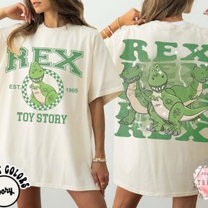May include: Ivory-colored t-shirt featuring a green graphic of the character Rex from Toy Story. The front design includes the word "REX" above a cartoon dinosaur in a circle, with "EST. 1995" and "TOY STORY" below. The back has a larger Rex graphic.