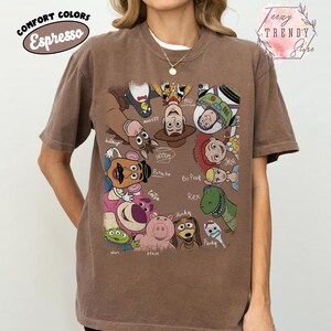 May include: Espresso-colored t-shirt featuring a colorful graphic of characters from the Toy Story movie franchise. The design includes Woody, Buzz Lightyear, and other characters. The shirt has the words "COMFORT COLORS" and "Espresso" printed on it.