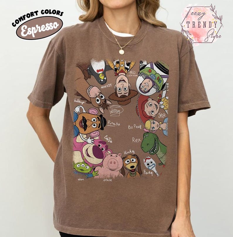 May include: Espresso-colored t-shirt featuring a colorful graphic of characters from the Toy Story movie franchise. The design includes Woody, Buzz Lightyear, and other characters. The shirt has the words "COMFORT COLORS" and "Espresso" printed on it.