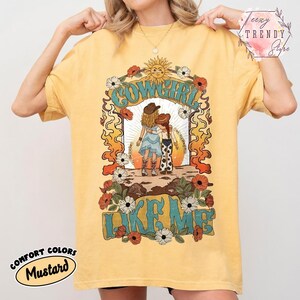 May include: A mustard yellow t-shirt featuring a graphic print of two figures in cowboy hats and boots standing together. The text COWGIRL LIKE ME is printed in large blue letters. The design includes a sun, flowers, and orange flame patterns.