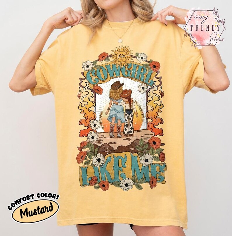 May include: A mustard yellow t-shirt featuring a graphic print of two figures in cowboy hats and boots standing together. The text COWGIRL LIKE ME is printed in large blue letters. The design includes a sun, flowers, and orange flame patterns.