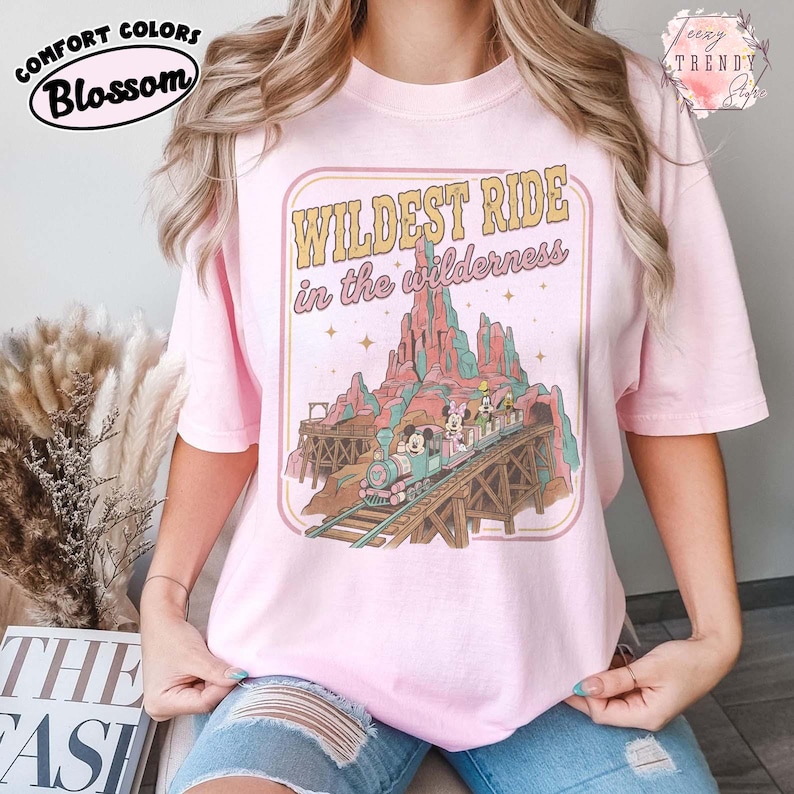 Może przedstawiać: R&oacute;żowa koszulka z grafiką pociągu na drewnianym moście, g&oacute;rami i napisem "Wildest Ride in the wilderness". Koszulka ma logo Comfort Colors Blossom w lewym g&oacute;rnym rogu.