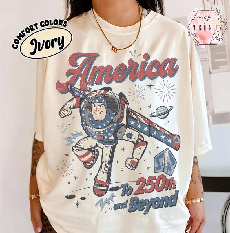 Puede incluir: Una camiseta de color marfil con un gr&aacute;fico de un personaje de ranger espacial con un traje rojo, blanco y azul. El texto dice America, To 250th and Beyond, acompa&ntilde;ado de ilustraciones de fuegos artificiales, estrellas, un planeta y un cohete.