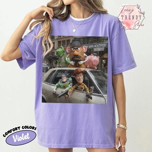 Puede incluir: Una camiseta violeta de manga corta con un estampado gr&aacute;fico de cinco personajes de juguete de una pel&iacute;cula. Los personajes incluyen un dinosaurio verde, una figura con forma de patata, un cerdo rosa, un ranger espacial y un vaquero con armas.