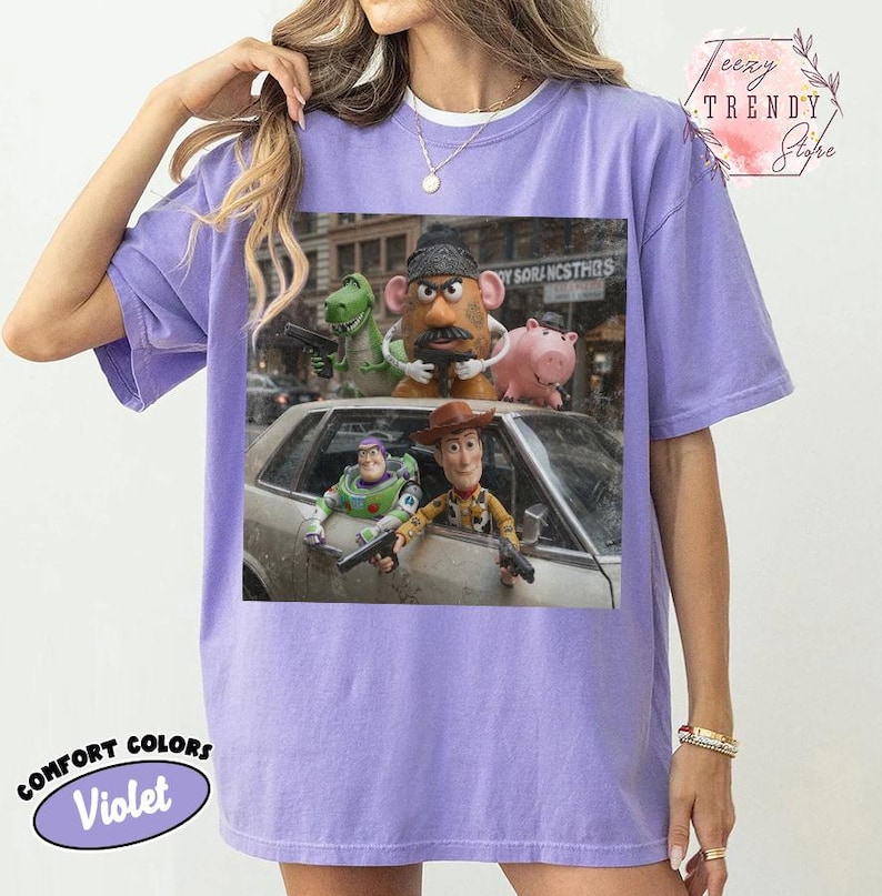 Puede incluir: Una camiseta violeta de manga corta con un estampado gr&aacute;fico de cinco personajes de juguete de una pel&iacute;cula. Los personajes incluyen un dinosaurio verde, una figura con forma de patata, un cerdo rosa, un ranger espacial y un vaquero con armas.