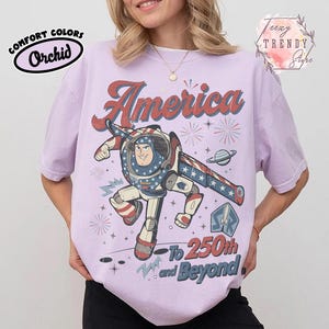 Puede incluir: Una camiseta de manga corta de color morado claro con un gr&aacute;fico de un personaje de ranger espacial con un traje blanco, rojo y azul. El texto dice America en la parte superior y To 250th and Beyond en la inferior, junto a estrellas, fuegos artificiales y un planeta.