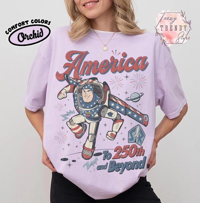 Puede incluir: Una camiseta de manga corta de color morado claro con un gr&aacute;fico de un personaje de ranger espacial con un traje blanco, rojo y azul. El texto dice America en la parte superior y To 250th and Beyond en la inferior, junto a estrellas, fuegos artificiales y un planeta.