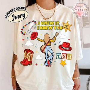 May include: An ivory t-shirt featuring a graphic print with the text "I KNEW IT, I KNEW YOU" and "INFINITY AND BEYOND". The design includes a person playing a guitar, a beaded bracelet, red cowboy boots, a red hat, and blocks labelled "TS5".