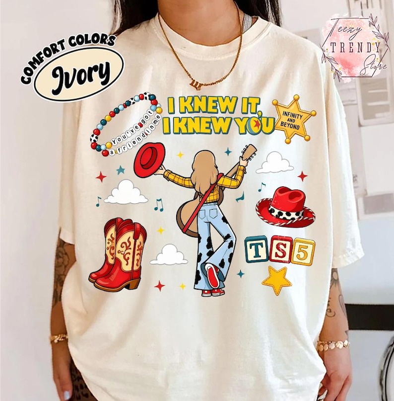 May include: An ivory t-shirt featuring a graphic print with the text "I KNEW IT, I KNEW YOU" and "INFINITY AND BEYOND". The design includes a person playing a guitar, a beaded bracelet, red cowboy boots, a red hat, and blocks labelled "TS5".