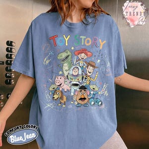 May include: Blue jean colored t-shirt featuring the words "TOY STORY" in colorful letters. The shirt has a graphic of characters from the Toy Story movie. The shirt also has the words "COMFORT COLORS Blue Jean" printed on the bottom.