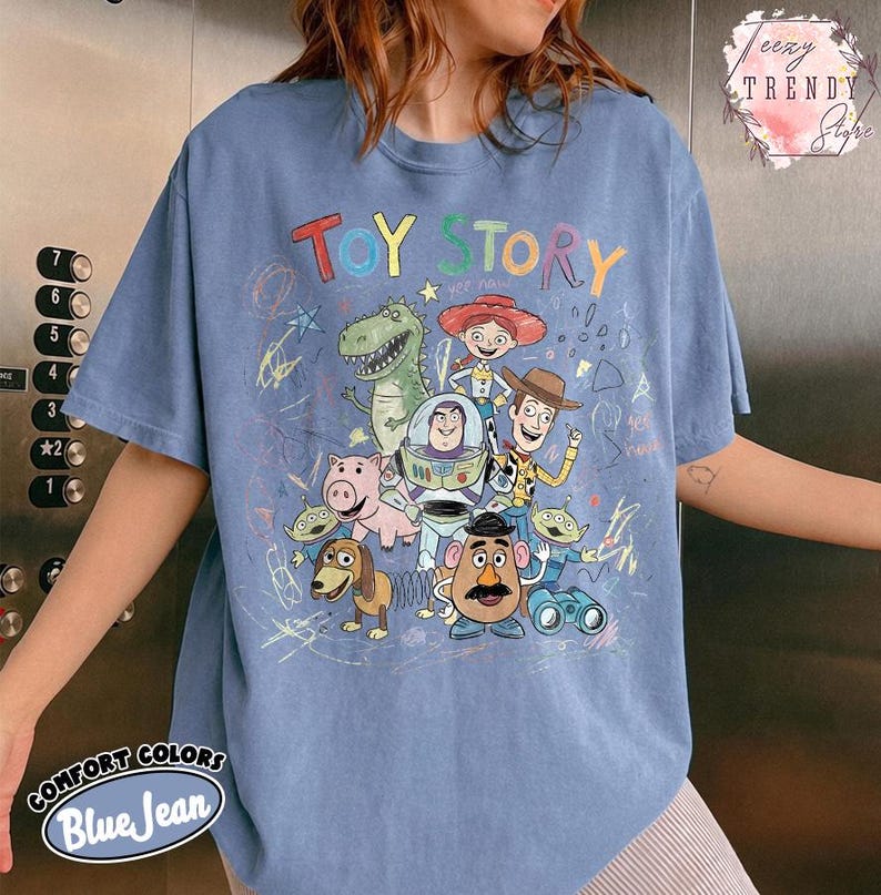 May include: Blue jean colored t-shirt featuring the words "TOY STORY" in colorful letters. The shirt has a graphic of characters from the Toy Story movie. The shirt also has the words "COMFORT COLORS Blue Jean" printed on the bottom.
