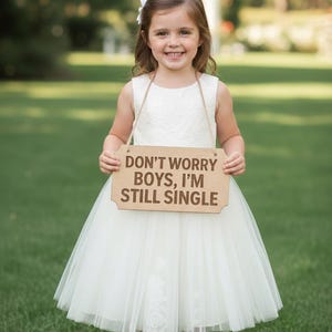 May include: A young girl in a white dress holds a wooden sign that reads "DON'T WORRY BOYS, I'M STILL SINGLE." The dress has a lace bodice and a tulle skirt. She is standing on green grass.