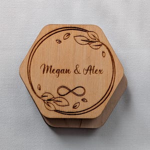 May include: A wooden, hexagonal box with a hinged lid. The lid is engraved with the names "Megan & Alex" inside a circular design with leaves and an infinity symbol. The box is light brown.
