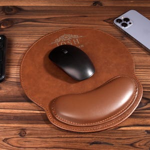 May include: A brown leather mouse pad with a wrist rest and the text "Josh" in gold lettering. A black wireless mouse is on the pad.