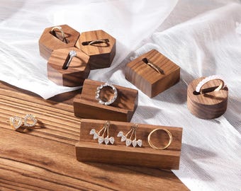Può includere: Una collezione di porta anelli in legno di varie forme, che espongono anelli e orecchini. I porta anelli sono realizzati in legno scuro e presentano fessure e scanalature per contenere gioielli. Alcuni anelli hanno pietre trasparenti e alcuni orecchini hanno un design pendente.