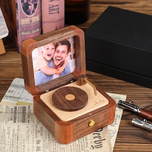 K&ouml;nnte beinhalten: Eine Holz-Spieluhr in Form eines Vintage-Plattenspielers mit einem Foto eines Mannes und eines Kindes. Die Box besteht aus dunklem und hellem Holz, mit einem goldfarbenen Tonarm und einem kleinen Knopf. Das Foto ist in der Box gerahmt.