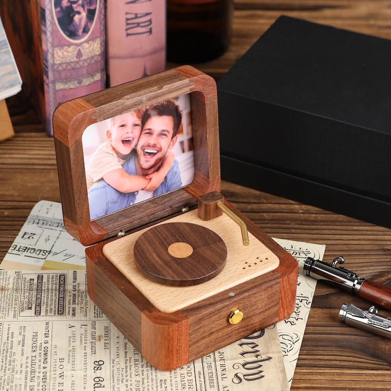 K&ouml;nnte beinhalten: Eine Holz-Spieluhr in Form eines Vintage-Plattenspielers mit einem Foto eines Mannes und eines Kindes. Die Box besteht aus dunklem und hellem Holz, mit einem goldfarbenen Tonarm und einem kleinen Knopf. Das Foto ist in der Box gerahmt.