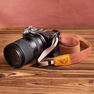 May include: A silver Sony Alpha 7C camera with a black lens and a tan and orange camera strap. The strap has a yellow leather patch with the initials "LR" and a deer antler design. The camera rests on a wooden surface.