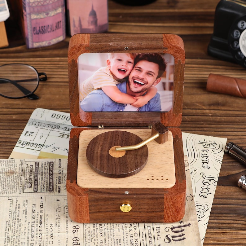 K&ouml;nnte beinhalten: Eine Holz-Musikbox mit einem Fotorahmen, der einen Mann und ein Kind zeigt. Die Box hat ein Plattenspieler-Design mit einem Arm in Messingoptik. Die Box ist aus dunklem Holz mit hellem Holz im Inneren und einer kleinen Schublade.