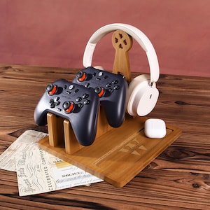 May include: A wooden gaming accessory organizer with a dark wood finish. It holds two black game controllers with orange accents, white headphones, and a white charging case. The organizer has a decorative wooden stand.