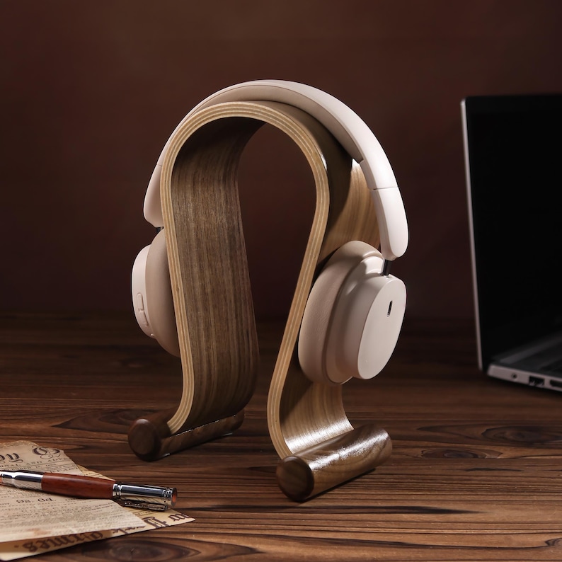 Puede incluir: Un soporte para auriculares de madera con un acabado marr&oacute;n claro. El soporte tiene forma de auricular y tiene una base curva. Un par de auriculares blancos est&aacute;n apoyados en el soporte.