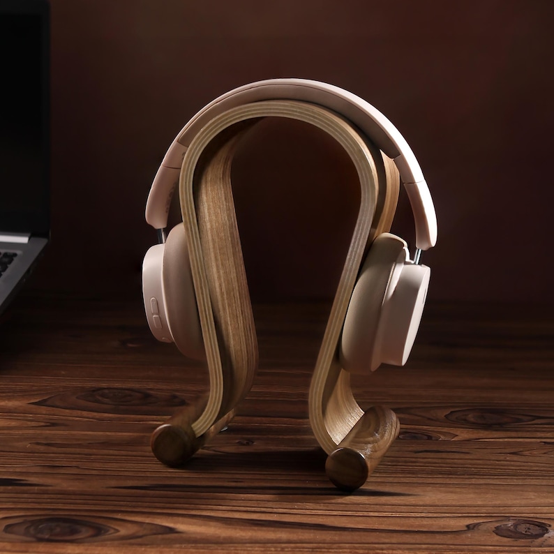 Puede incluir: Un soporte para auriculares de madera con un dise&ntilde;o curvo. El soporte est&aacute; hecho de madera marr&oacute;n claro y tiene un acabado natural. Un par de auriculares de color rosa claro est&aacute;n apoyados en el soporte.
