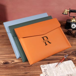 May include: Three leather document holders in blue, green, and orange. The orange holder has a flap closure and features the letter "R" with the name "ROBERT" printed below. The holders are arranged on a wooden surface.