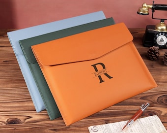 May include: Three leather document holders in blue, green, and orange. The orange holder has a flap closure and features the letter "R" with the name "ROBERT" printed below. The holders are arranged on a wooden surface.