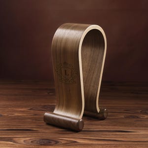 Puede incluir: Un soporte para auriculares de madera con un dise&ntilde;o curvo. El soporte est&aacute; hecho de madera de nogal y tiene un acabado marr&oacute;n claro. El soporte tiene un peque&ntilde;o logotipo grabado en la parte delantera que dice "Luxury".