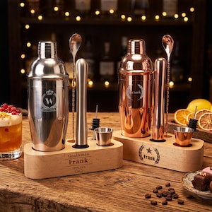 May include: Two cocktail shaker sets on light wood stands. The left set is silver with a W crest and a stand engraved with "Gentleman Frank 1999.12.1". The right set is copper with a laurel wreath crest and a stand engraved with "Frank". Each includes a muddler, spoon, pourer, and jigger.