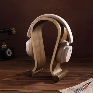 Puede incluir: Un soporte para auriculares de madera con un dise&ntilde;o curvo. El soporte est&aacute; hecho de madera marr&oacute;n claro y tiene un acabado natural. Un par de auriculares blancos est&aacute;n apoyados en el soporte.