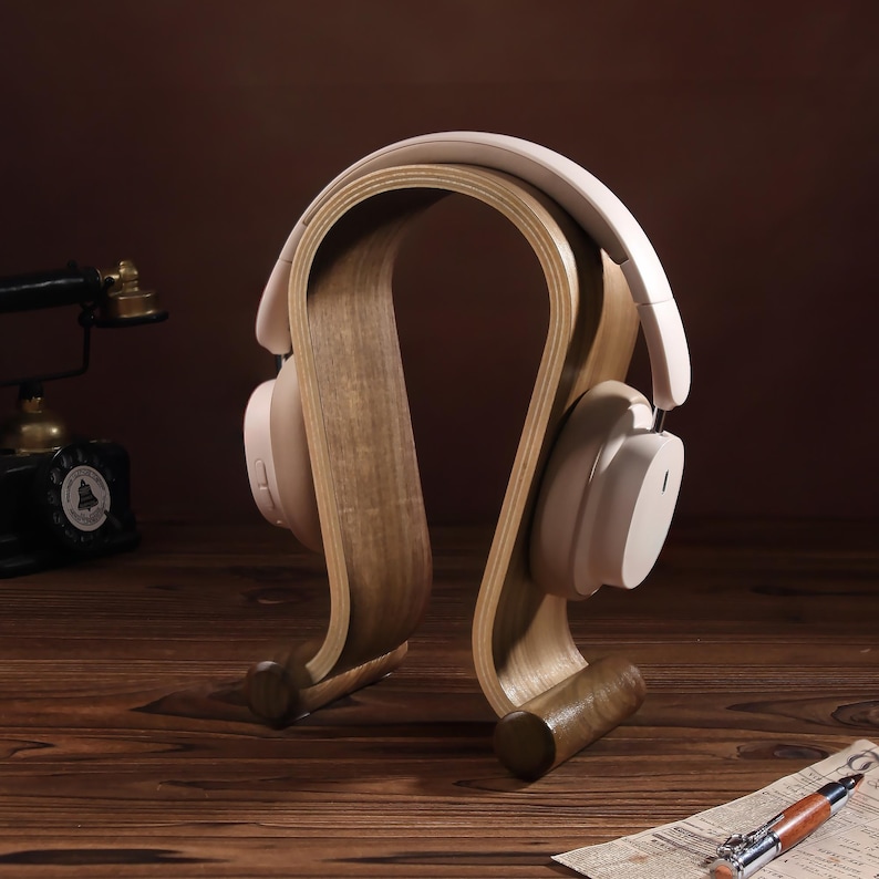 Puede incluir: Un soporte para auriculares de madera con un dise&ntilde;o curvo. El soporte est&aacute; hecho de madera marr&oacute;n claro y tiene un acabado natural. Un par de auriculares blancos est&aacute;n apoyados en el soporte.
