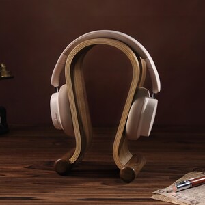 Puede incluir: Un soporte para auriculares de madera con un dise&ntilde;o curvo. El soporte est&aacute; hecho de madera marr&oacute;n claro y tiene un acabado natural. Un par de auriculares blancos est&aacute;n apoyados en el soporte.