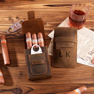 Puede incluir: Un estuche de cuero marrón para puros con tres puros y un cortapuros. El estuche está abierto, revelando los puros y el cortapuros. También hay un estuche cerrado con las iniciales "LK", junto con un vaso de líquido ámbar y puros sobre una superficie de madera.