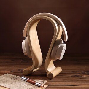 Puede incluir: Un soporte para auriculares blanco de madera con un dise&ntilde;o curvo. El soporte sostiene un par de auriculares blancos.